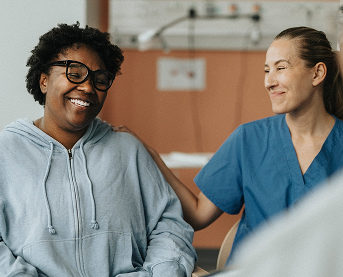 Woman in gray sweater smiling while comforted by woman in blue scrubs